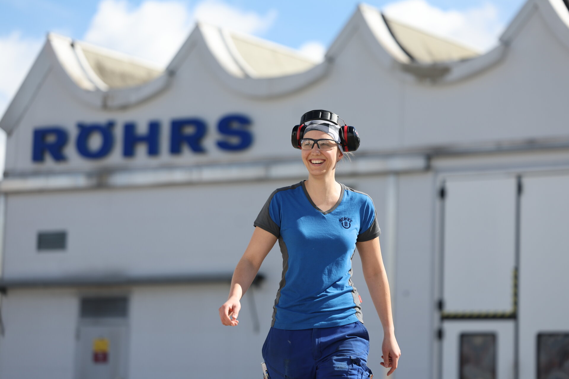 Röhrs Mitarbeiterin mit Gehörschutz läuft lachend vor der Röhrs Produktionshalle